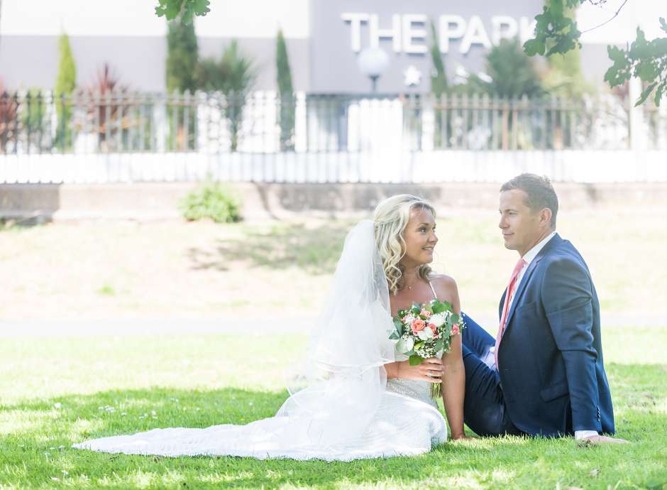 Bride and groom sat in the grass with the park hotel in background